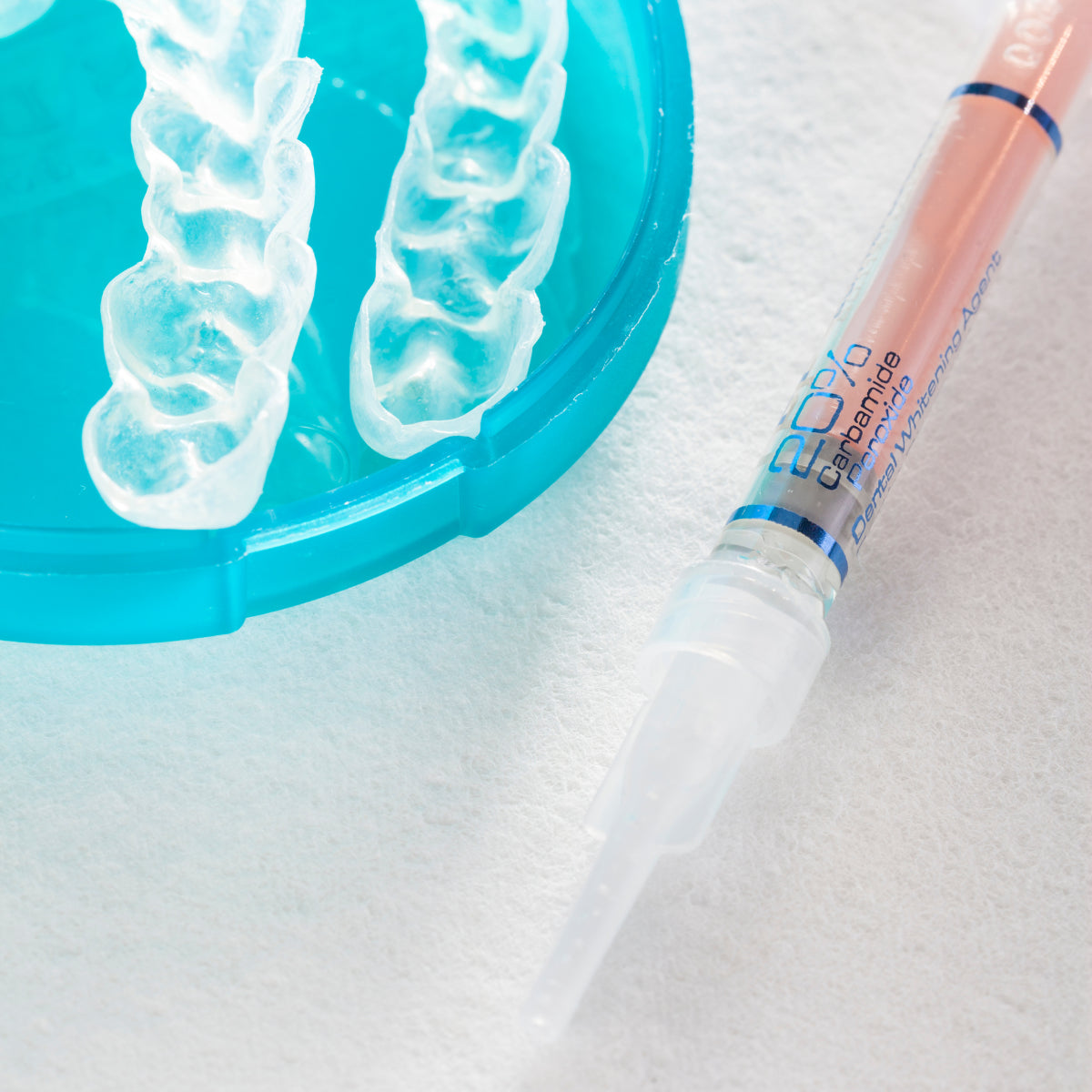 Striekačka s bieliacim gélom na zuby vedľa priehľadného zubného náustku na modrej podložke.